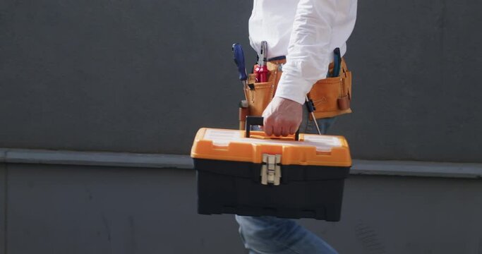 Builder confidently walks across site wearing tool-belt carrying helmet and sturdy toolbox. Serviceman ready to tackle daily tasks