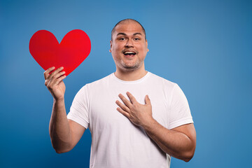 man holding a paper heart in his right hand and his left hand touching his chest, surprised