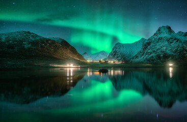 Aurora borealis, snowy mountains, sea, fjord, reflection in water, street lights at starry winter night. Lofoten, Norway. Northern lights. Landscape with polar lights, snowy rocks, sky with stars - obrazy, fototapety, plakaty