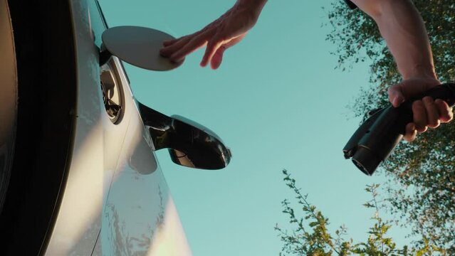 Male hand disconnects power connector from EV car, low angle, closeup. Unrecognizable man unplug charging cable from electric vehicle and finishes charge on background of blue sky and green trees
