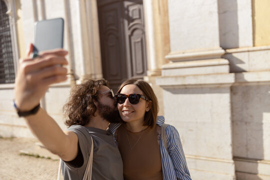 Happy tourist couple walking in city street, kissing and making selfie on phone. High quality photo