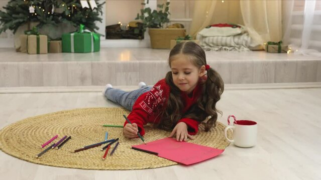Cute happy little girl lying on the floor in living room and writing a letter to Santa Claus. Happy New Year holidays