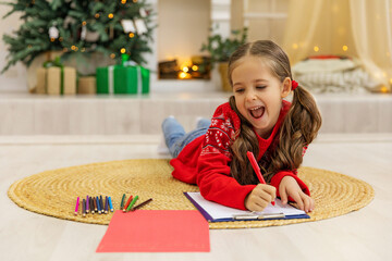Cute happy little girl lying on the floor in living room and writing a letter to Santa Claus. Happy...