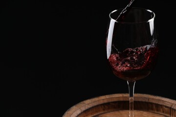 Pouring red wine into glass on wooden barrel against black background, closeup. Space for text