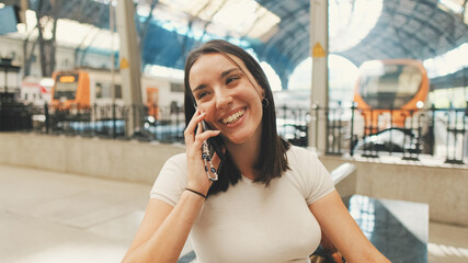 Happy beautiful girl traveler sits on the platform of the railway station, talking on the mobile...