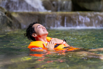 Young Asian man travel nature on summer holiday vacation. Happy generation z guy in life vest relax and enjoy outdoor lifestyle jumping and playing water at waterfall lagoon in forest mountain.