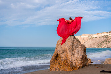 woman sea red dress. Woman with long hair on a sunny seashore in a red flowing dress, back view, silk fabric waving in the wind. Against the backdrop of the blue sky and mountains on the seashore.