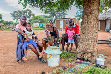 village african family, sited in the yard in front of the house, hut with thatched roof in the...
