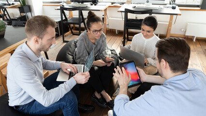Focused multiracial colleagues sit in circle in office work on modern gadgets during casual meeting. Concentrated diverse multiethnic employees busy with devices at briefing at workplace.