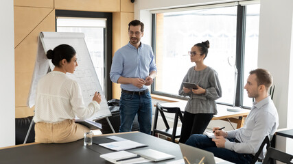 Female coach make flip chart presentation for diverse employee at office training or meeting. Multiracial businesspeople discuss whiteboard presentation in group at workplace. Teamwork concept.
