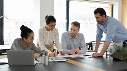 Happy multiethnic colleagues coworkers gather at desk in office discuss company financial paperwork together. Smiling multiracial businesspeople brainstorm collaborate with documents at workplace.