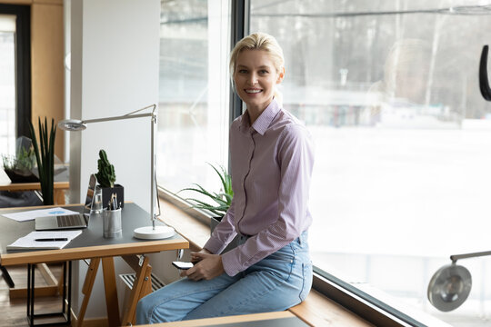 Portrait of smiling young Caucasian female employee pose in modern workplace with smartphone. Happy woman worker in office, show confidence and leadership and success. Business concept.