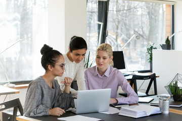 Concentrated multiethnic female colleagues look at laptop screen brainstorm cooperate in office. Focused multiracial women work together on computer, discuss company business project in group.