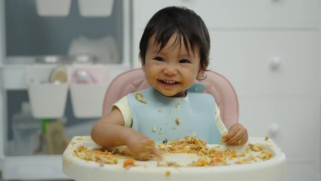 happy infant baby eating food and vegetable by self feeding BLW or baby led weaning on a chair