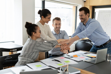 Happy multiethnic colleagues stack join hands engaged in teambuilding activity or training at workplace. Overjoyed multiracial employees have fun feel motivated from shared success or achievement.