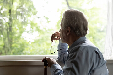 Retirement problem. Pensive old man nursing home patient sit by window hold glasses walking stick...