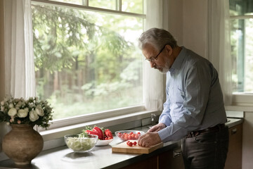 Aged man cooking. Inspired mature grey haired grandpa cook morning breakfast at home kitchen alone. Concentrated mature male prepare salad cut fresh vegetables on table at country cottage. Copy space