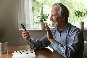 Videocall. Happy grey haired aged man grandpa using phone to talk to grandchild online wave hand to...