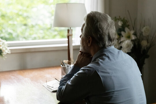 Far away in thoughts. Rear view of aged hoary male sit by table at home alone look at window ponder prop chin on folded hands. Thoughtful mature man going down to memories reviewing past. Copy space - Powered by Adobe
