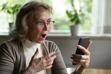 Shocked with phone message. Stunned elderly woman in glasses look on cellphone screen with opened...