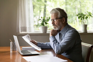 Engaged in paperwork. Serious aged businessman working from home office sit at desk read loan contract letter correspondence. Retired hoary man in glasses think on financial papers study bank report - obrazy, fototapety, plakaty