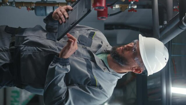 Professional heavy industry worker wearing safety uniform and hard hat works on tablet computer. Male inspector checks piping system on modern factory or industrial energy facility. Vertical view.