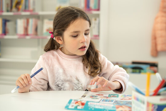 Little caucasian girl in pink sweater and with curly hair at a child library painting and reading. High quality photo