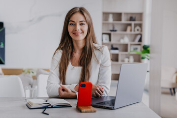 Purposeful young cheerful woman in business attire sitting at desk with laptop phone, notebook looks at camera ready for remote video conference at home. Home office.