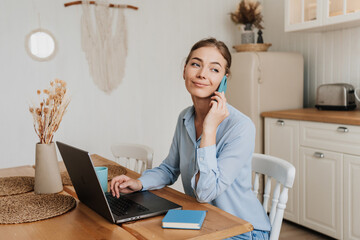 Grateful blonde girl in casual sitting at desk wit laptop talks by phone looks away, remote working young woman at home. Home office. - obrazy, fototapety, plakaty