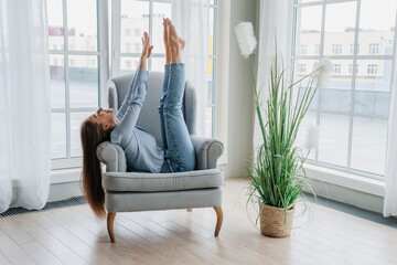 Relaxed caucasian girl in jeans sitting in cozy chair having fun rising hands and legs, doing nothing at home, reboot, weekend. Caress student enjoying vacations. - obrazy, fototapety, plakaty
