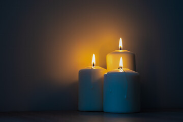 Three burning candles in the dark. Warm light from candles against cold dark background. - obrazy, fototapety, plakaty