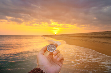 hand holds a compass against the background of the sea. the setting sun falls on the beach and...