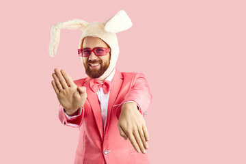 Funny extravagant man in pink suit and hat with rabbit ears dancing on pink background. Caucasian man in red glasses in formal suit and humorous hat makes funny movements looking at camera. Banner.