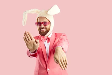 Gordijnen Funny extravagant man in pink suit and hat with rabbit ears dancing on pink background. Caucasian man in red glasses in formal suit and humorous hat makes funny movements looking at camera. Banner. © Studio Romantic