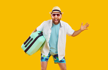 Happy male tourist with suitcase. Joyful bearded man in summer clothes, cool sunglasses and fedora sunhat holding his bag and standing isolated on yellow color background. Travel, holiday concept - obrazy, fototapety, plakaty
