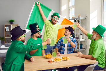 Group of male friends celebrating St. Patrick day with Irish flag. Cheerful bearded men in carnival Leprechaun hats toasting with green beer drinks and having fun indoors