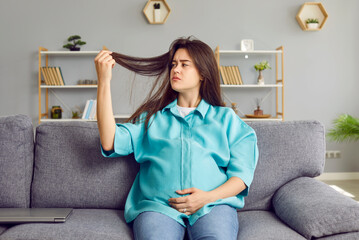 Portrait of a young brunette sad pregnant woman in casual clothes sitting on sofa in the living room at home and looking on her long hair falling out. Pregnancy and hair loss problems. - obrazy, fototapety, plakaty