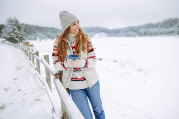 Beautiful young woman drinks tea with a thermos. Great winter weather, snowy lake. Winter walk. Young female tourist enjoying the weather outdoors.