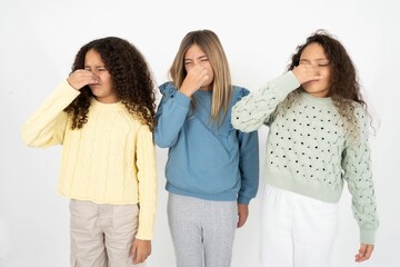 Three beautiful multiracial kid girls  smelling something stinky and disgusting, intolerable smell, holding breath with fingers on nose. Bad smell - obrazy, fototapety, plakaty