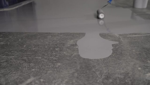 Painting the floor with a roller. Repair or construction work. Close-up of a paint roller applying a new coat of gray paint to the floor, the plinth protected with yellow masking tape.