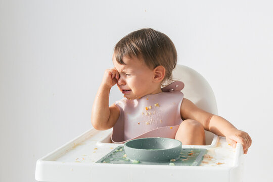 cute crying baby sitting eating lunch in high chair at home on white background, hysterics and tears in a 1 year and 3 month old child