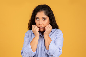 A worried young woman with curly hair wearing a blue shirt bites her nails and looks directly at the camera