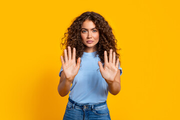 Curly-haired woman gesturing stop with hands on yellow background - obrazy, fototapety, plakaty