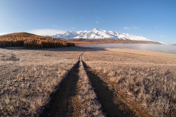 Remote dirt trail through the frozen steppe to snow mountains. Bright sunny autumn atmospheric...