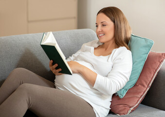 Smiling pregnant woman in casual clothes reading book at home