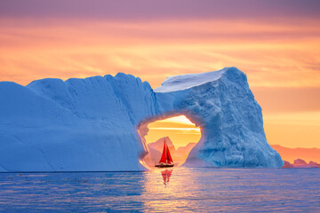 Beautiful landscape with little red sailboat cruising under a majestic iceberg arch during midnight sun season of polar summer in Greenland. Sail boat with red sails cruising among icebergs, Greenland