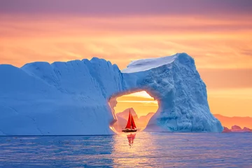 Foto op Canvas Beautiful landscape with little red sailboat cruising under a majestic iceberg arch during midnight sun season of polar summer in Greenland. Sail boat with red sails cruising among icebergs, Greenland © Revive Photo Media