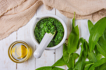 Fresh bear garlic also known as wild garlic or ursinum allium pesto crushed in a mortar and oil on a wooden table with white boards and beige kitchen table cloth. Top view.