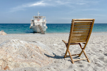 Empty wooden chair on sand beach faces towards boat and sea panorama. Summer travel concept.