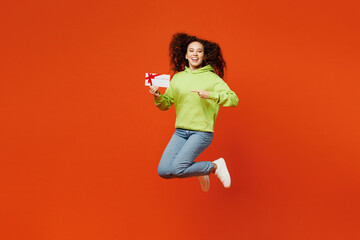 Full body young woman of African American ethnicity she wear green hoody casual clothes jump high hold point on gift certificate coupon voucher card for store isolated on plain red orange background.
